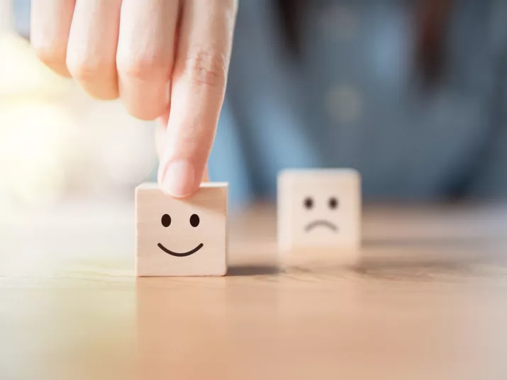 A hand touches a cube with a happy smiley face painted on it. A sad cube smiley can be seen in the background.